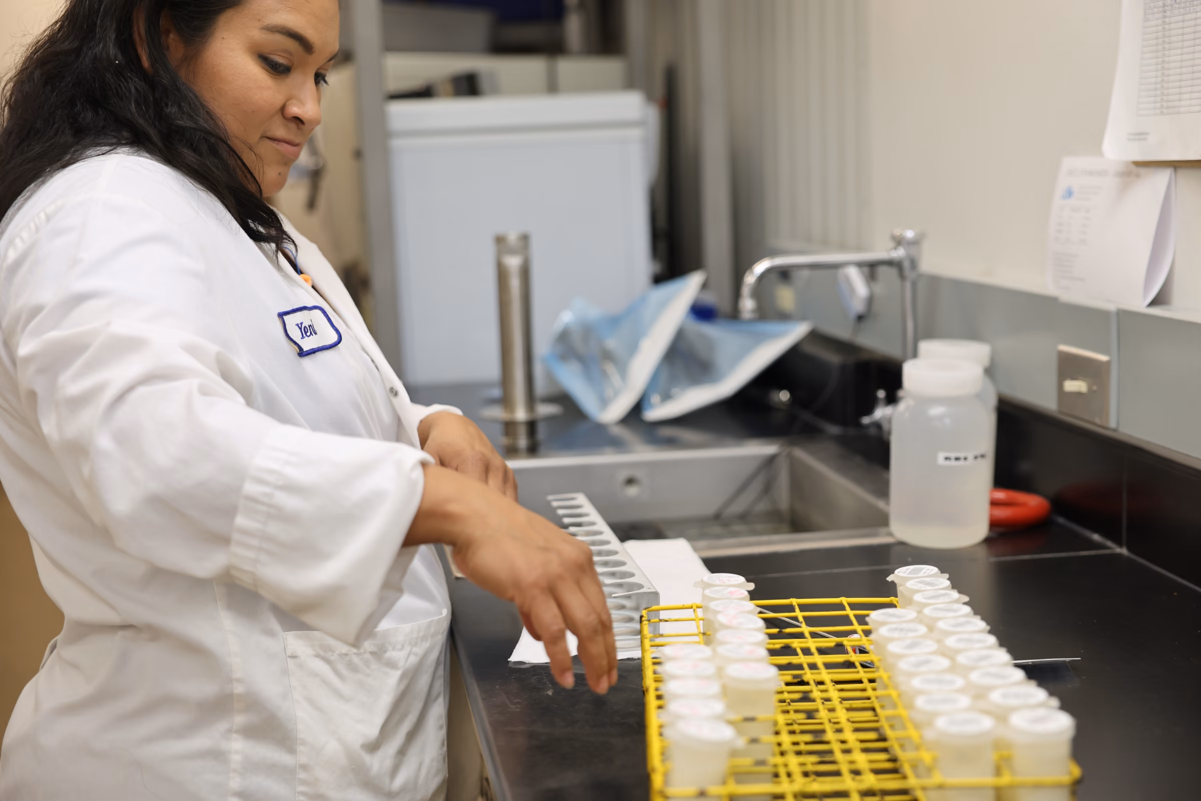 Woman in lab coat handling small containers in a lab with sink and equipment in background.
