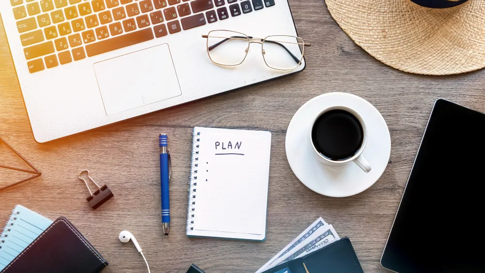 op-down view of a wooden desk with a laptop, eyeglasses, a cup of coffee, a notebook labeled ‘PLAN,’ a pen, and a smartphone.