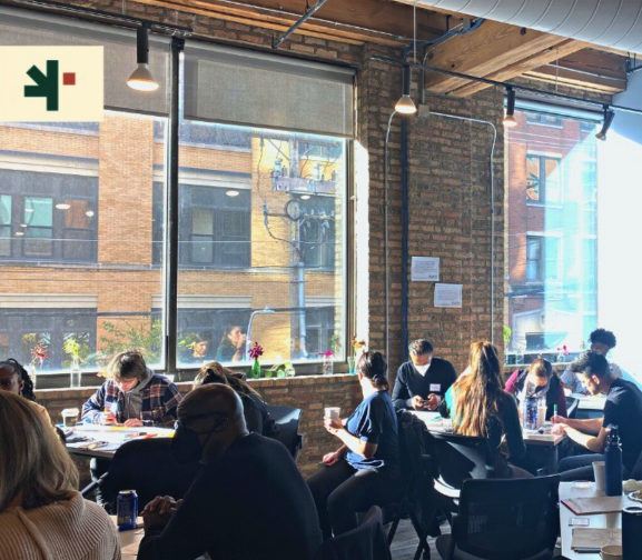 Groups of NewRoot staff members gathered around tables, some with heads down writing. They are in a room with large windows and exposed brick.