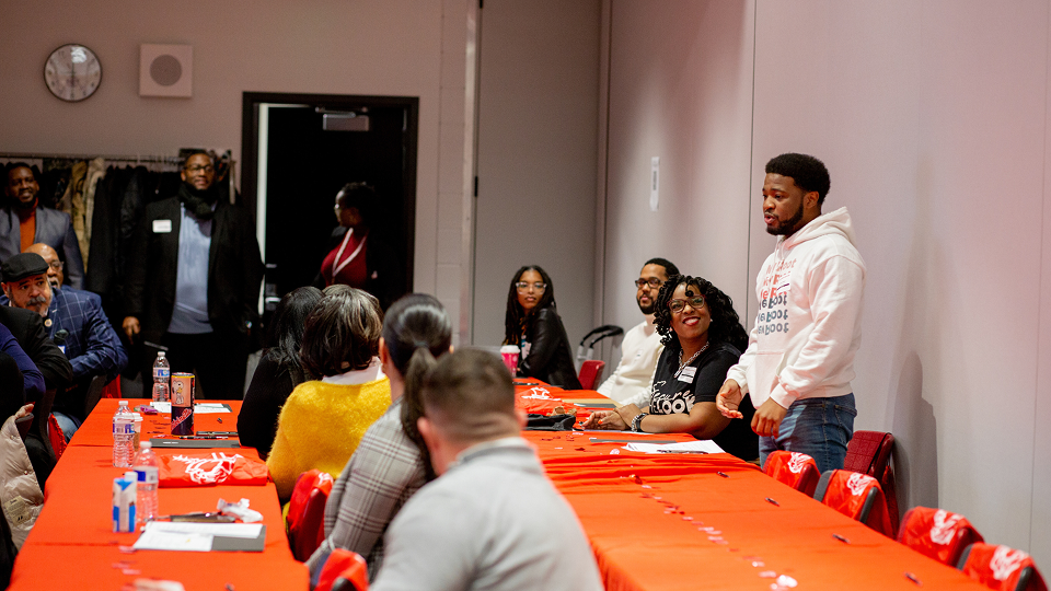 A group of adults is gathered around two long tables covered in red-orange tablecoths. One person wearing a white hoodie and jeans is addressing the group.