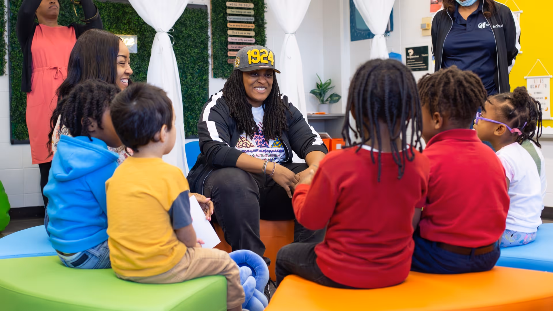 NewRoot facilitator, in black jacket and grey hat, is surrounded by a group of elementary school students as they participate in a Peace Circle. Adult educators observing outside of the circle.