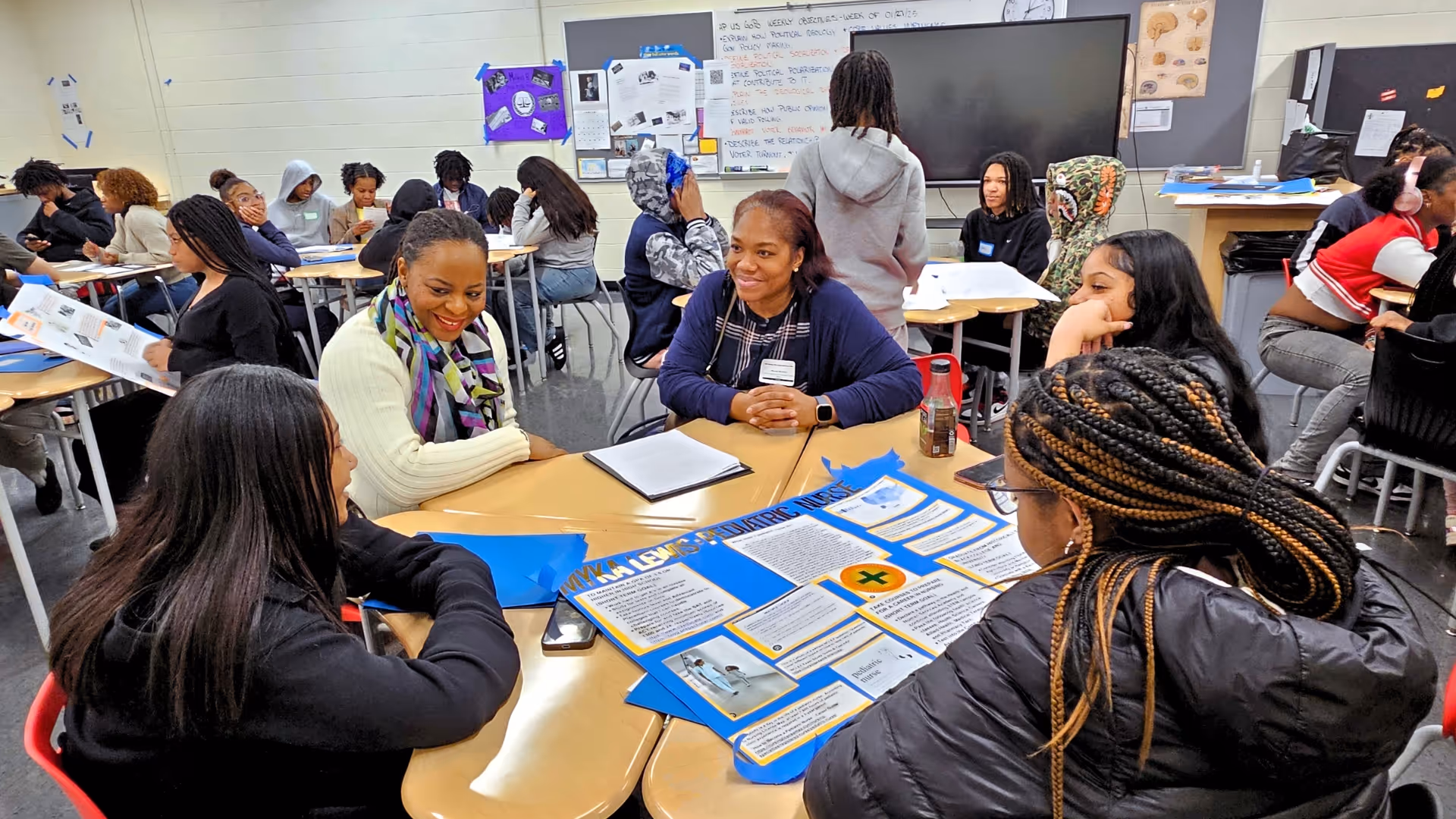 At one of NewRoot's Student Day of Action events, three high school students are seated with two adult volunteers, surrounding a vision board in a classroom.
