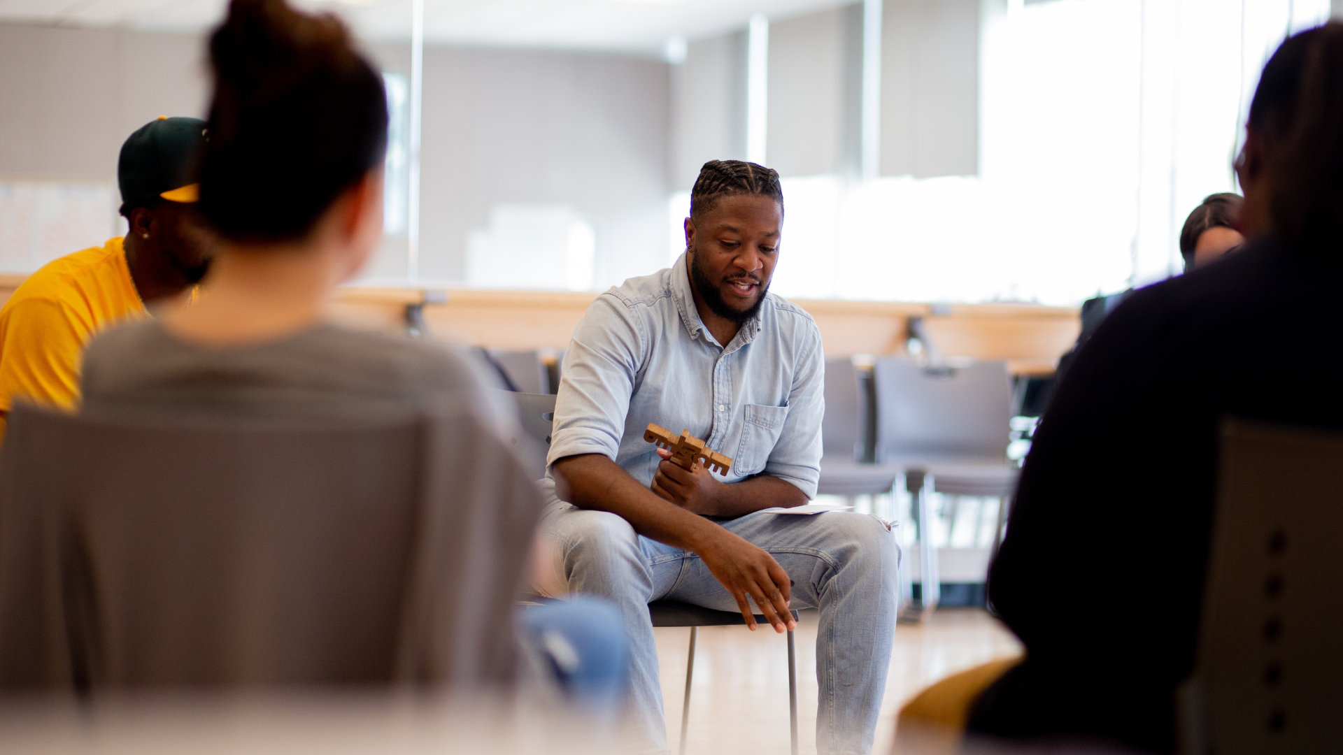 NewRoot facilitator, in denim shirt and jeans, is holding a talking piece, surrounded by adults seated in a circle.
