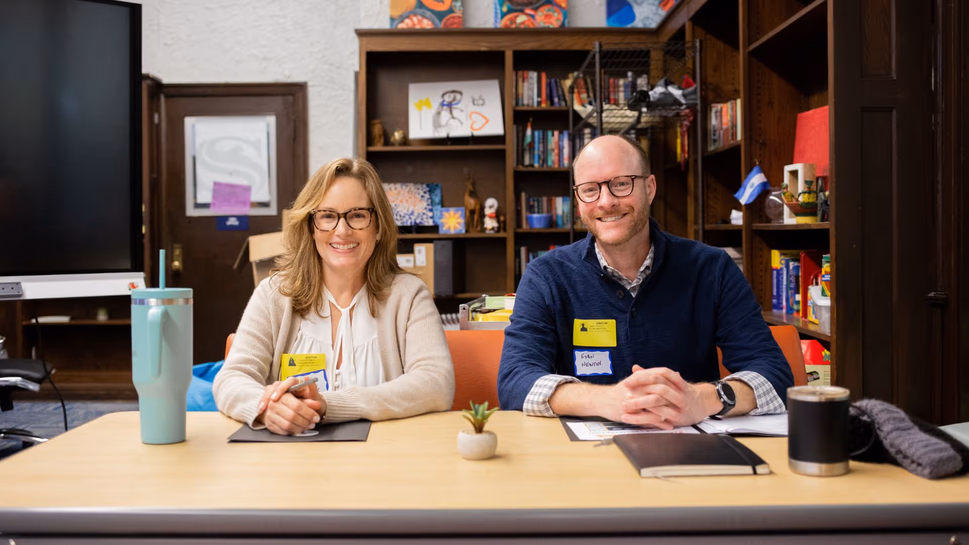 A man and woman sit at a table with books and a potted plant.