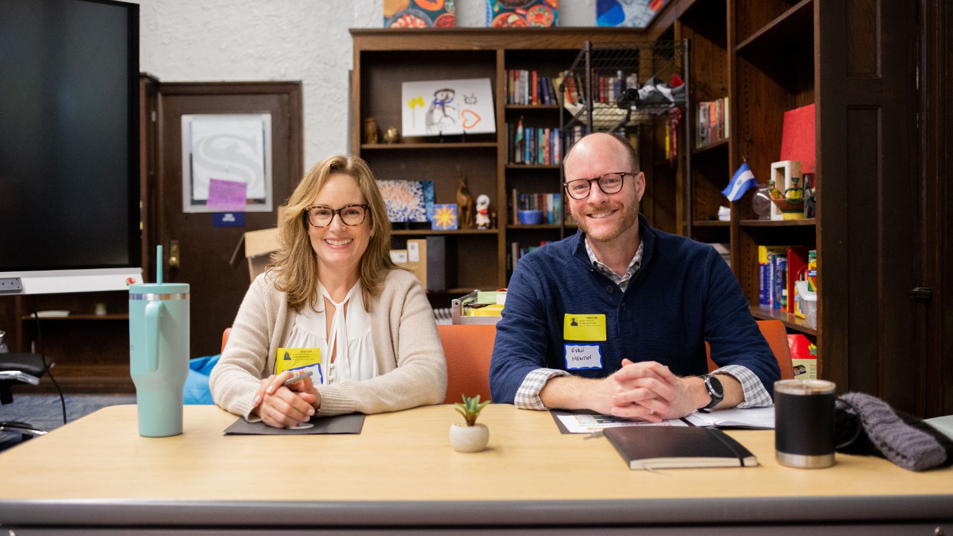 A man and woman sit at a table with books and a potted plant.