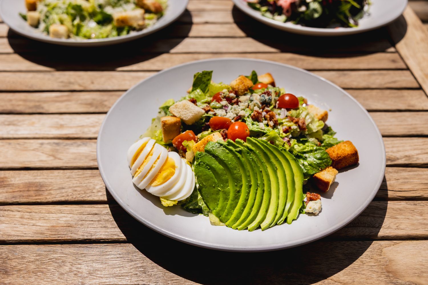 Plate with salad on it with avocado and tomatoes, two other plates of salad behind center salad