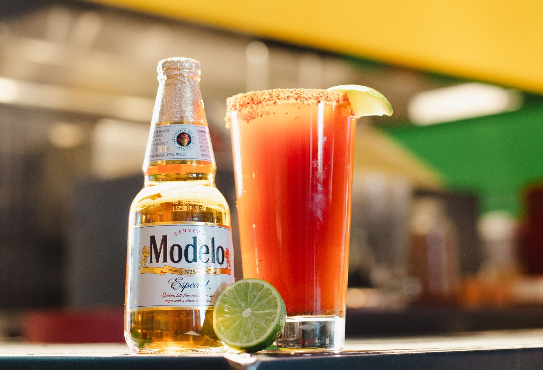 Bottle of Modelo with half a lime next to a glass of Michelada rimmed with Tajin and a lime wedge.