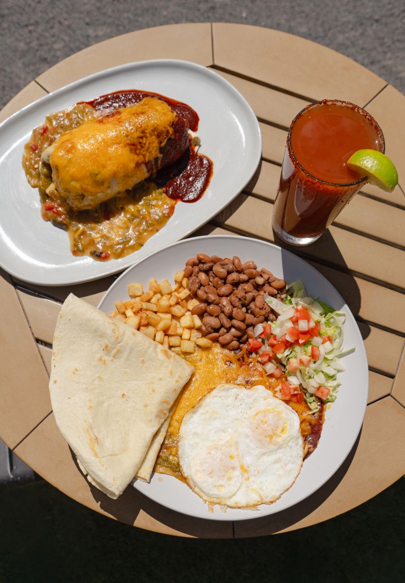 Glass of Michelada rimmed with Tajin and a lime wedge. Next to plate of Huevos Ranchers and red and green smothered breakfast burrito.