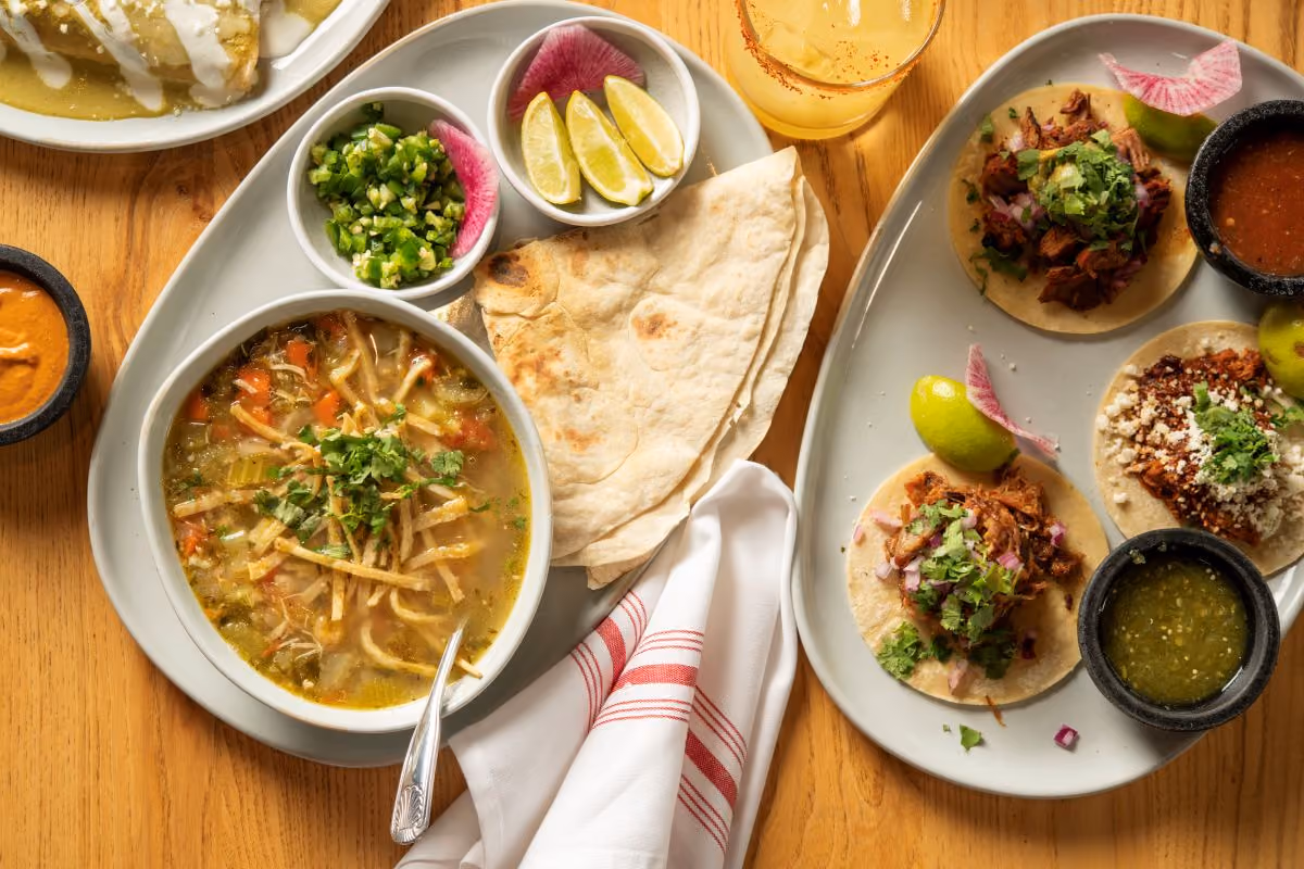 Overhead table view of Tortilla Stew with a side tortilla, also picture three tacos and sides of Salsa and crema