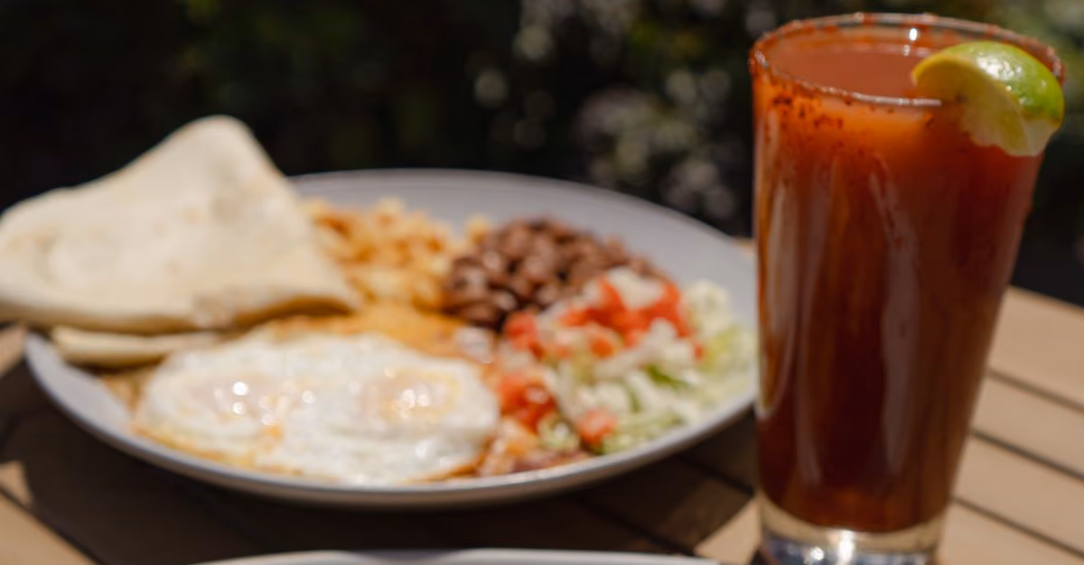Glass of Michelada rimmed with Tajin and a lime wedge. Next to plate of Huevos Ranchers and red and green smothered breakfast burrito. 