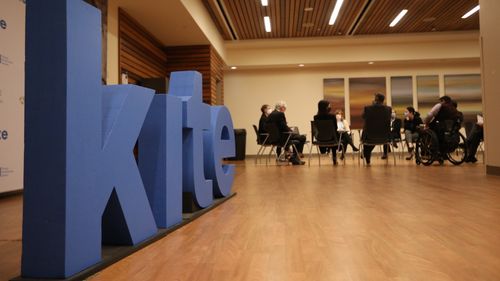 A "KITE" sign with a group of people meeting at Toronto Rehabilitation Institute's auditorium.