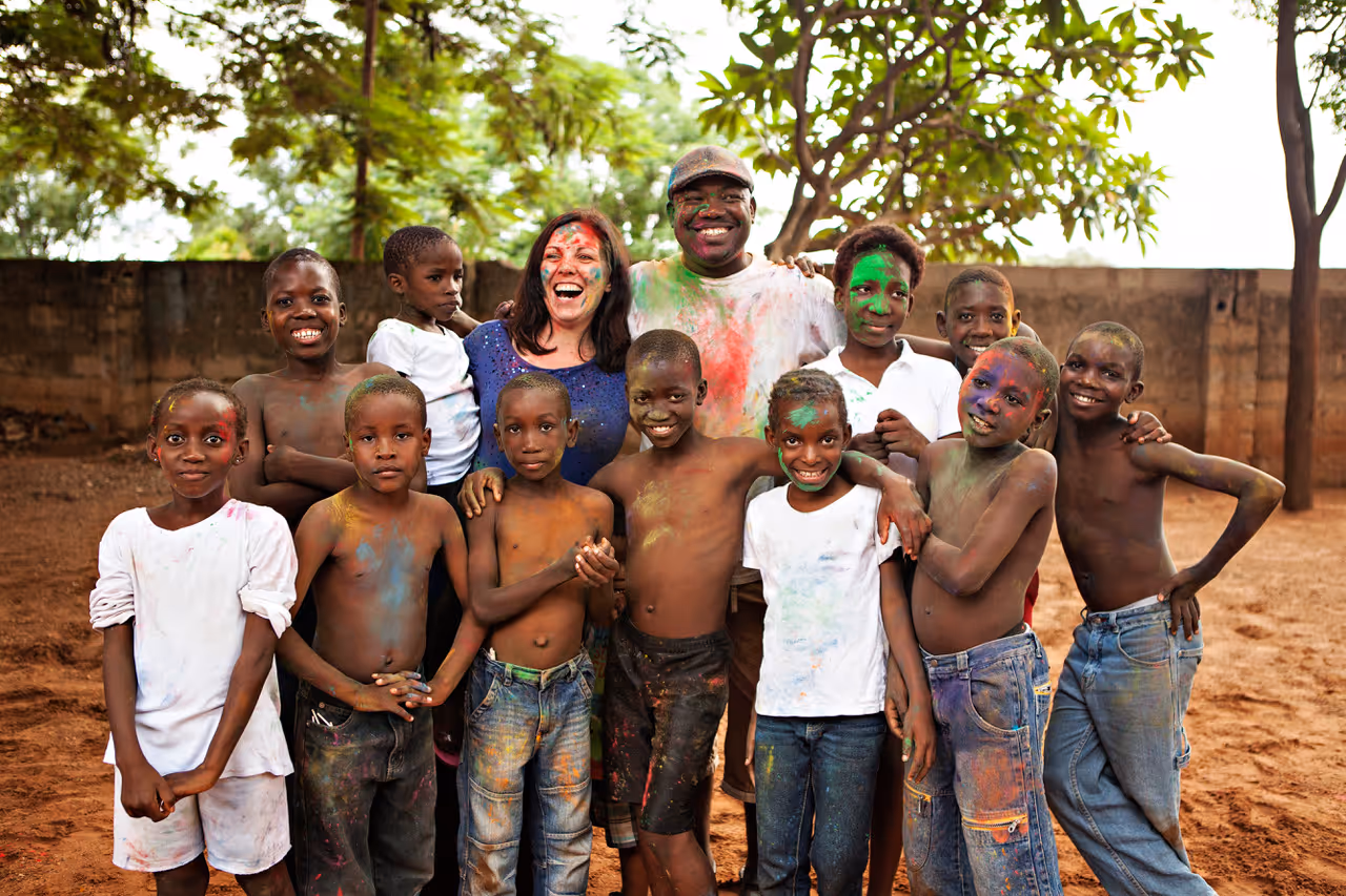 Jeanette und Agripa Phiri stehen lachend mit einer großen Gruppe von Kindern des Kwathu Children’s Home in Livingstone, Sambia, zusammen. Alle sind mit bunter Farbe bedeckt und posieren fröhlich im Freien unter Bäumen. Das Bild zeigt die enge, herzliche Gemeinschaft des Kinderheims.