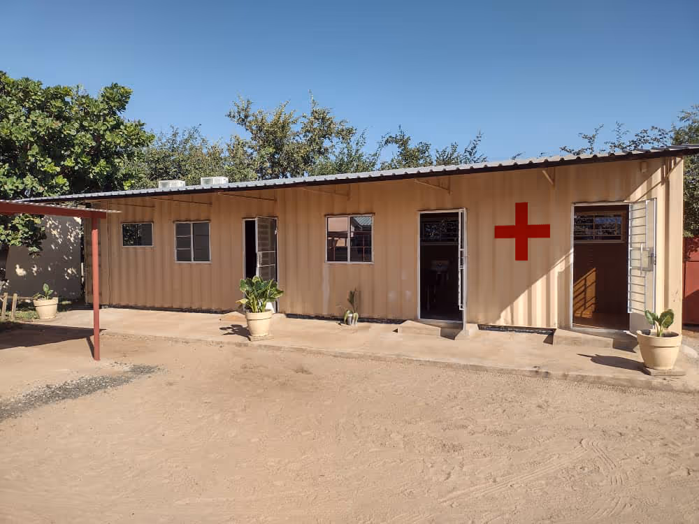 Ein einfacher Klinikbau aus Containern auf dem Gelände des Kwathu-Kinderheims in Livingstone, Sambia. Das lange, helle Gebäude mit Fenstern, offenen Türen und einem roten Kreuz an der Fassade steht auf einem sandigen Hof unter blauem Himmel.