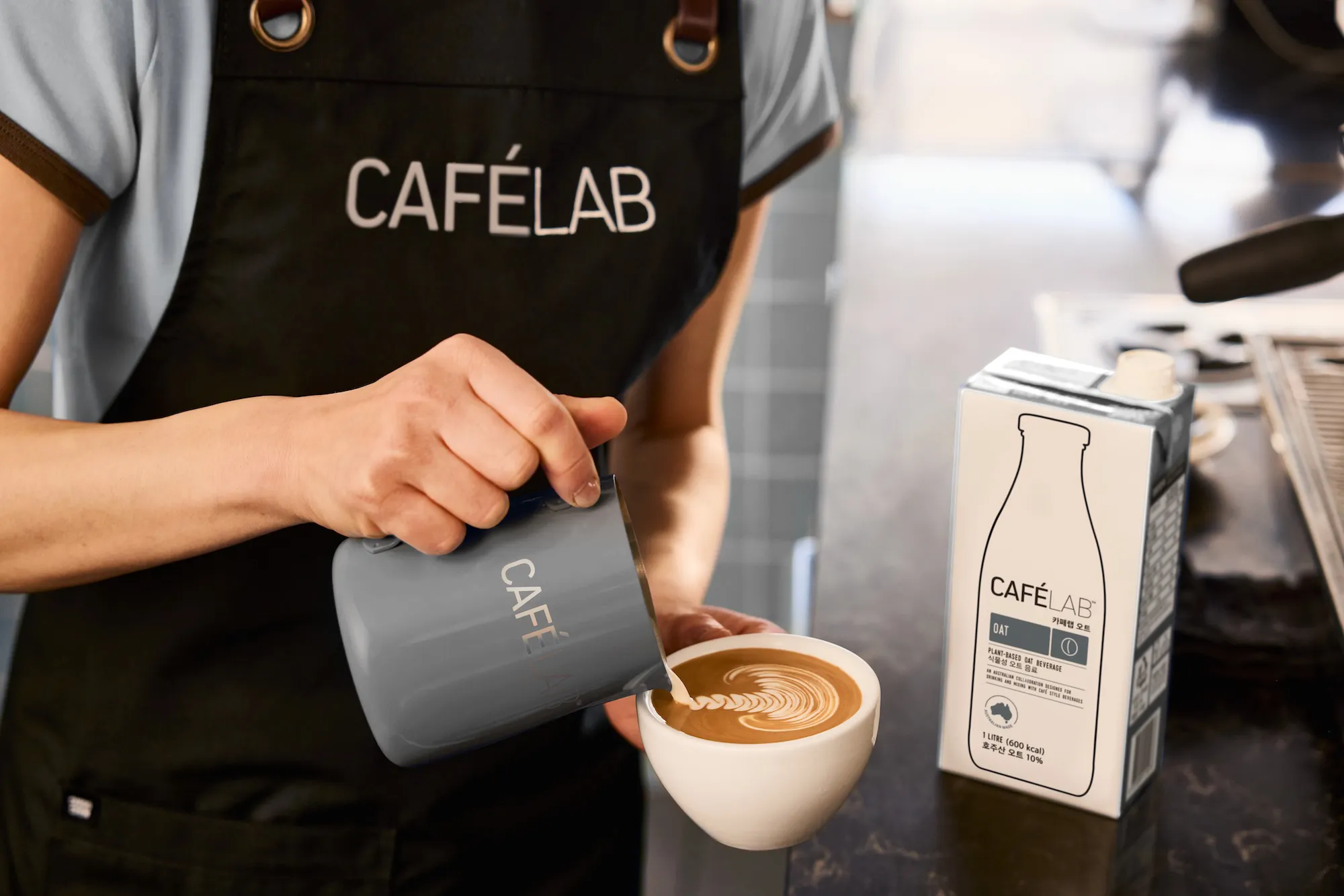 바리스타가 CAFÉLAB 로고가 새겨진 용기에서 라떼 아트를 위해 우유를 커피잔에 따르고 있음.