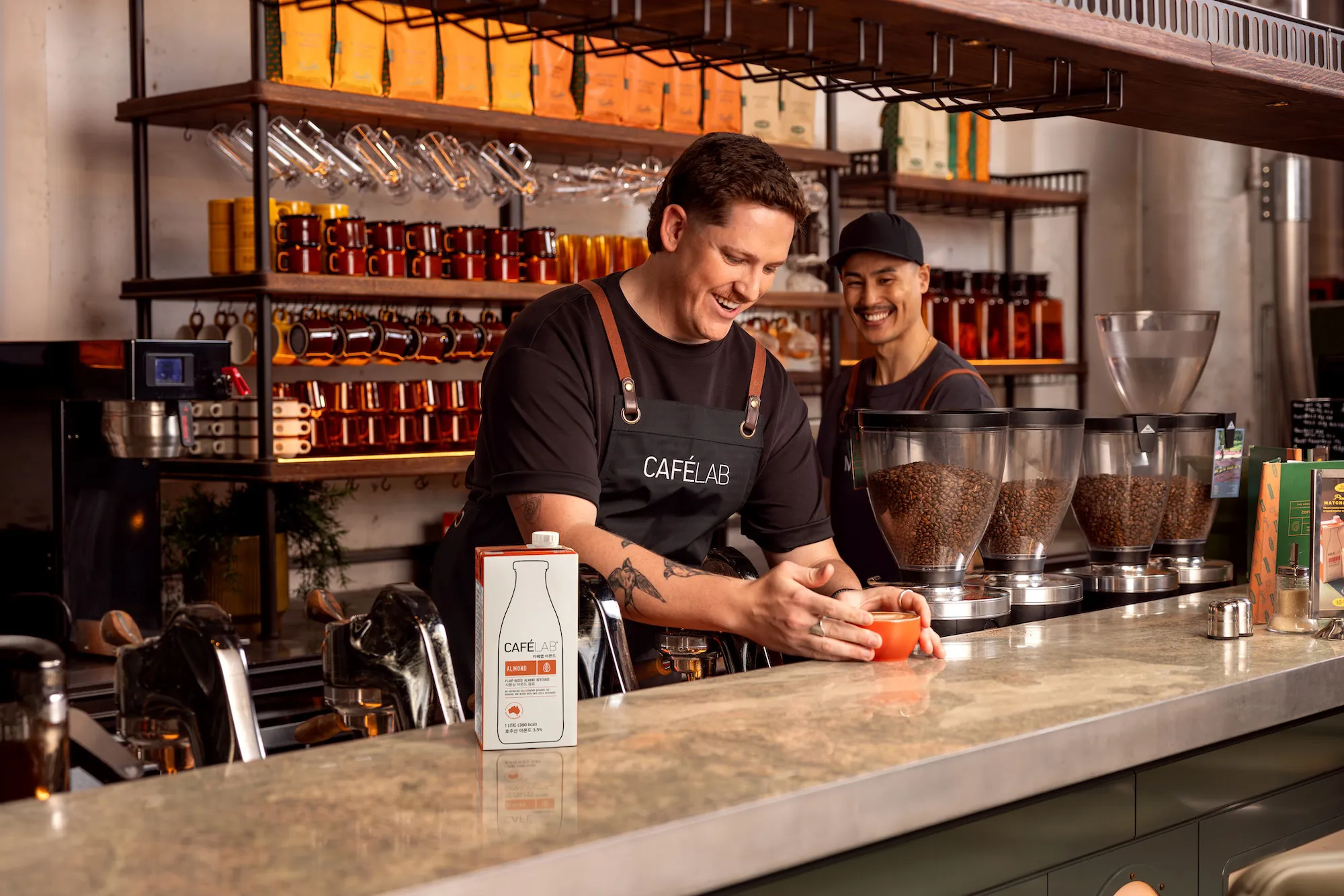 카페에서 커피잔을 준비하는 미소 짓는 바리스타 두 명과 원두가 담긴 큰 용기들이 놓인 카운터.