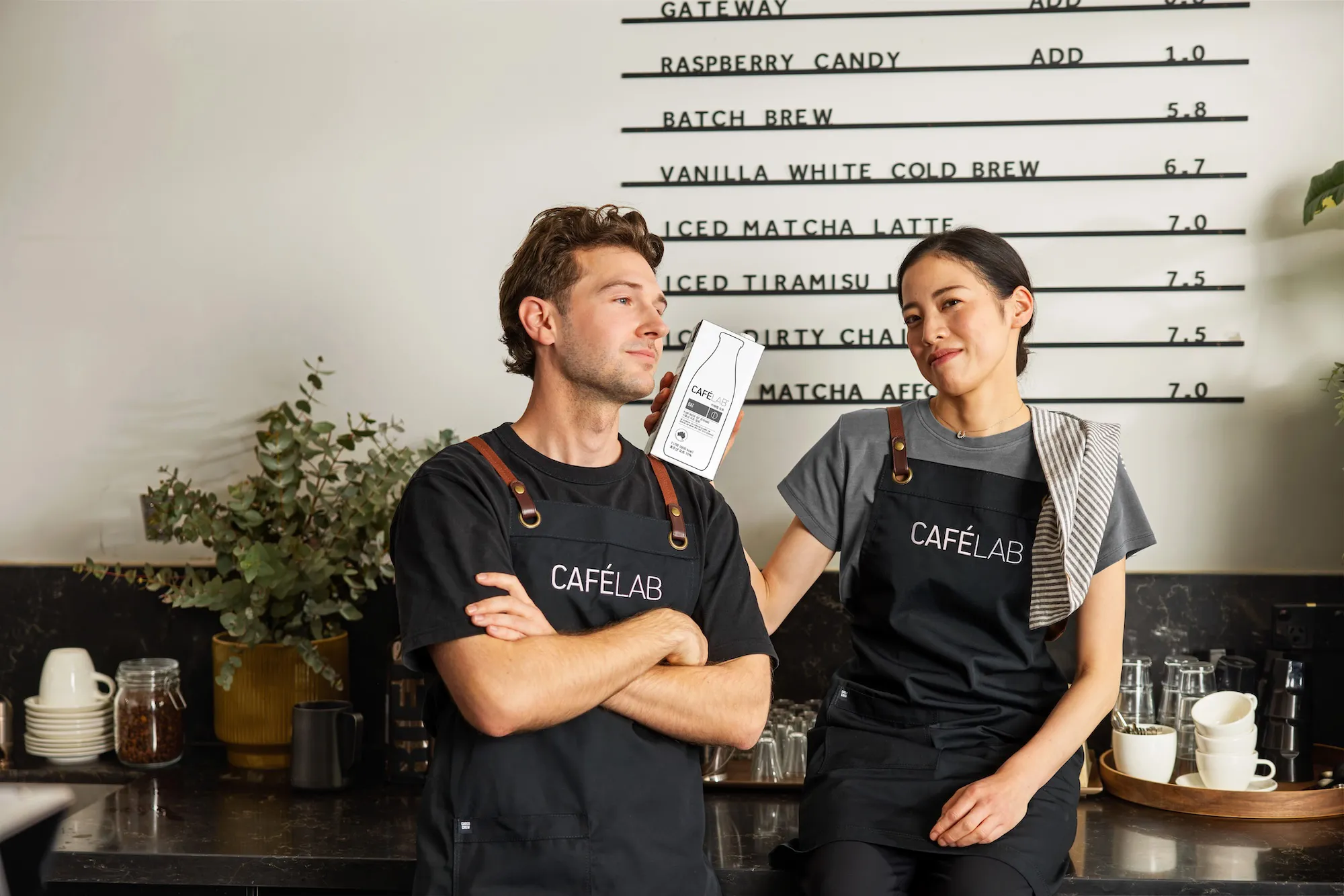카페 앞에서 검정색 CAFÉLAB 앞치마를 입은 남성과 여성이 함께 포즈를 취하고 있음.