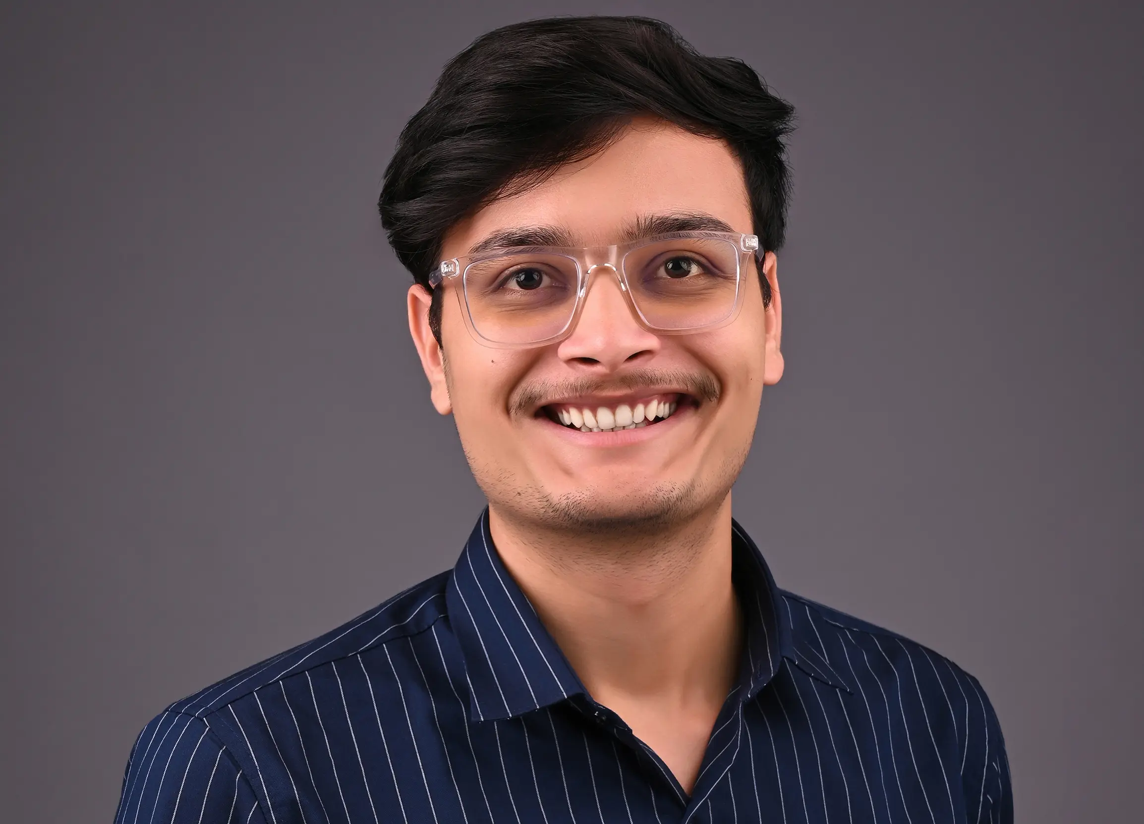 Headshot of Saksham Gupta;Photo of a young man in a striped dark blue shirt. He has glasses with thick transparent frame. He has black hair and thin mustache and stubble.