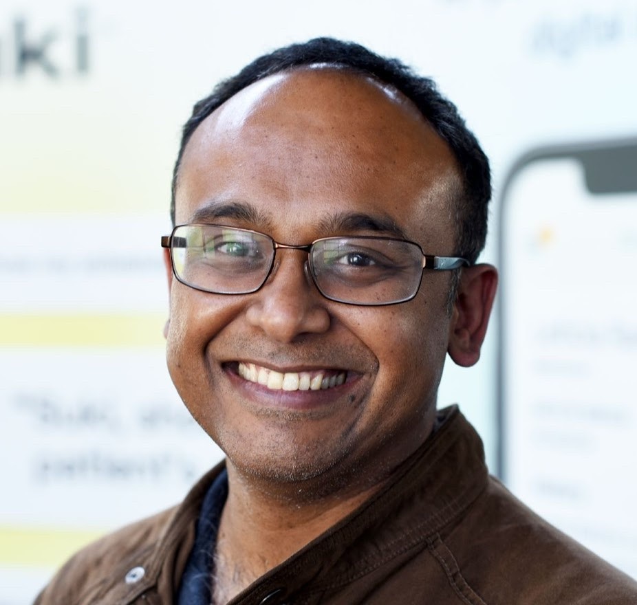 Headshot of Ajith Warrier; A smiling, bald man with dark hair on the sides, glasses, and a brown jacket. He is positioned in front of a blurred event backdrop.