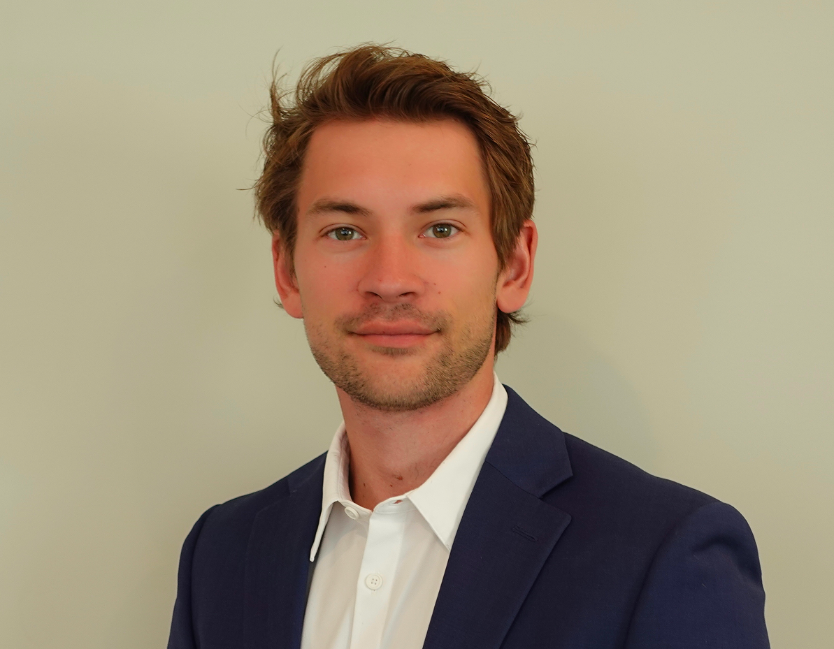 Headshot of Matthew Skaar;Photo of a young man in dark blue blazer and white shirt. He has brown hair and a small smile on his face. The background colour is grey.