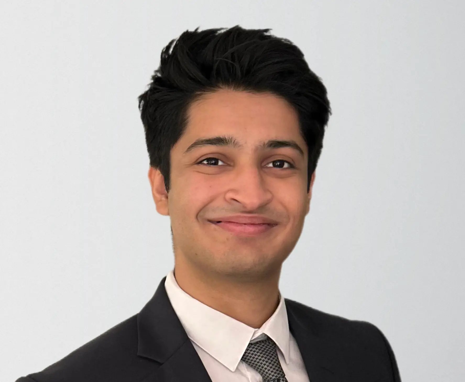 Headshot of Diwaker Sinha; A headshot of a young man with dark, styled hair smiling against a plain, light grey background. He is wearing a formal dark suit jacket over a white dress shirt and a grey textured tie.