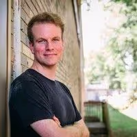 Photo of Brett Goodson; Portrait of a smiling man with short reddish-brown hair and crossed arms, wearing a dark t-shirt, standing outdoors with his back against a brick wall.