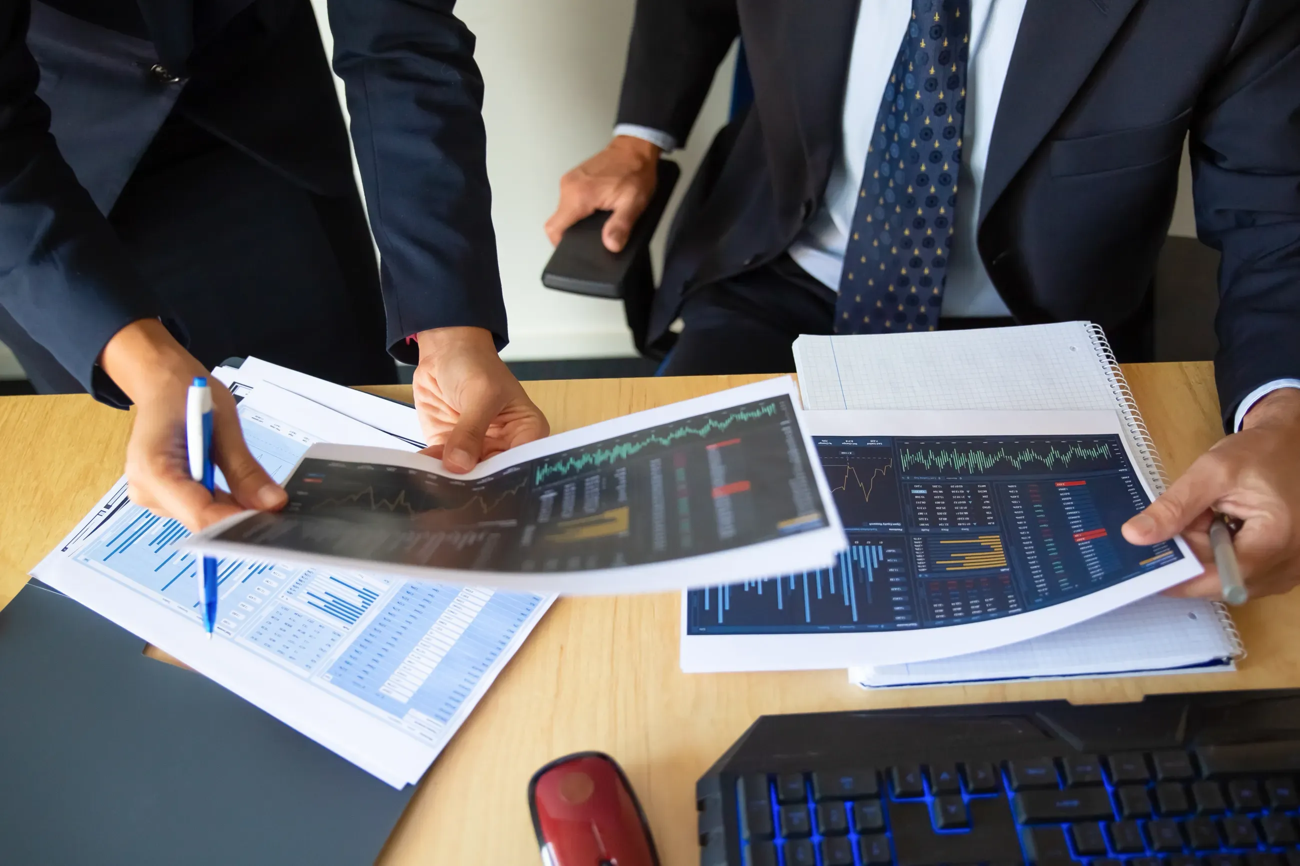 Graphic from the article Spacs Trended Up but Did Returns; Partial images of two people at an office desk. They are in formal suits and are comparing two images. Also seen on the desk are a keyboard, mouse, other printouts in a folder and a book. Both of the people also have a pen in their hands.
