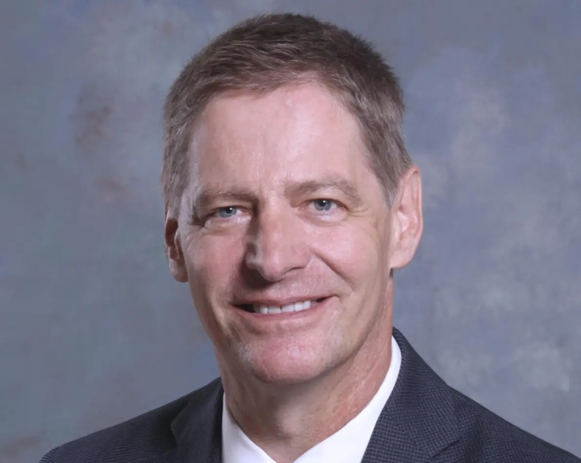 Photo of Lou Van Thaer; Headshot of a smiling man with short brown hair and blue eyes, wearing a dark suit jacket over a crisp white dress shirt against a mottled blue-grey studio background.