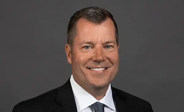 Photo of Mark; A headshot of a middle aged man wearing a black suit, white shirt, and patterned tie, photographed against a plain gray background.