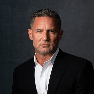 Photo of George Randall; Close-up portrait of a middle-aged man with short graying hair and light stubble, wearing a black suit jacket and an open-collared white shirt against a dark textured background.
