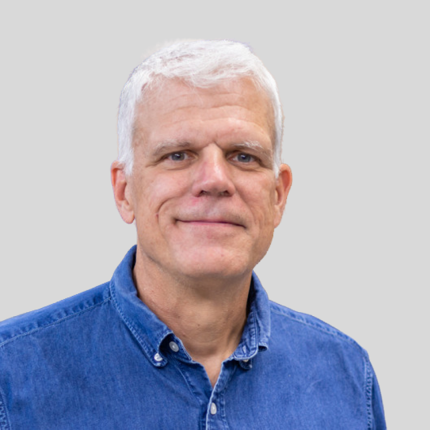 Photo of Glenn Daehn; Close-up portrait of an older man with short white hair and a neutral expression, wearing a blue denim button-down shirt against a plain light gray background.