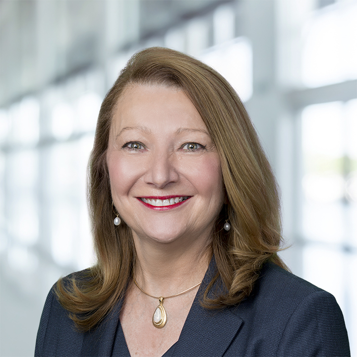 Photo of Suzanne Nimocks; Professional headshot of a smiling woman with brown hair, pearl earrings, and a gold pendant, wearing a dark blazer in a bright office.