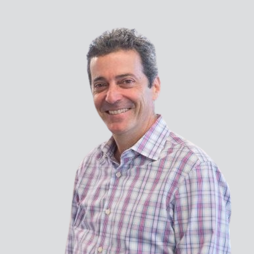 Photo of Paul Miller; Close-up portrait of a smiling man with short curly dark hair, wearing a white, blue, and purple plaid collared button-down shirt against a plain light gray background.