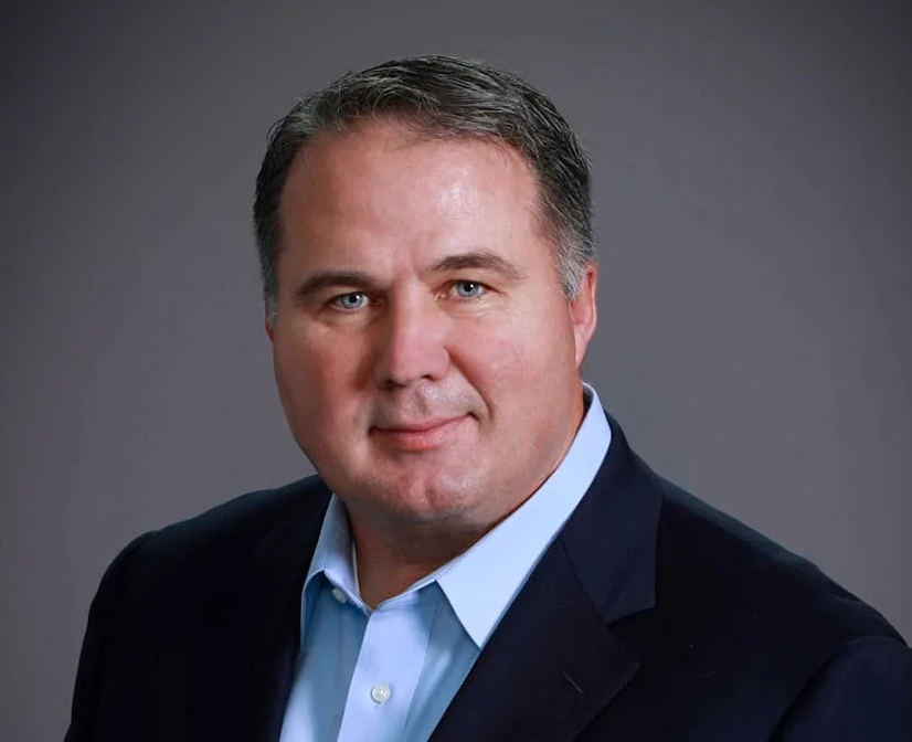 Photo of Bill J; Close-up portrait of a middle-aged man with short graying hair and a neutral expression, wearing a dark blue suit jacket and an open-collared light blue shirt against a plain gray background.