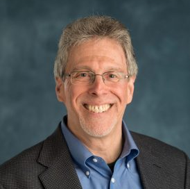 Photo of Mark; Close-up portrait of a smiling middle-aged man with short graying hair, thin-rimmed glasses, and light facial stubble, wearing a dark textured suit jacket over an open-collared blue shirt against a mottled blue-gray background.