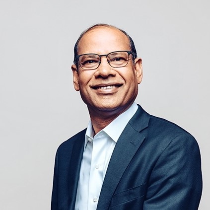 Photo of Anupam Khare; Close-up portrait of a smiling middle-aged man with short dark hair, dark-rimmed glasses, wearing a dark blue suit jacket over an open-collared white shirt against a plain light gray background.