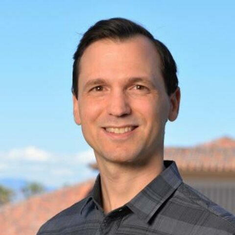 Photo of Michael Bernstein; A portrait of a smiling man with short brown hair, wearing a grey plaid collared shirt against a slightly blurred outdoor background featuring a clear blue sky and a terracotta-tiled roof.