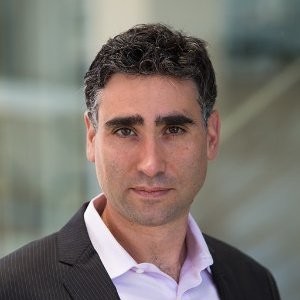 Photo of Martin Casado; A headshot of a man with short, wavy dark brown hair and a light beard stubble, looking directly into the camera with a neutral expression. He is wearing a dark pinstripe suit jacket over a light pink or lavender open-collar dress shirt. The background is softly blurred with a neutral, bright indoor lighting.