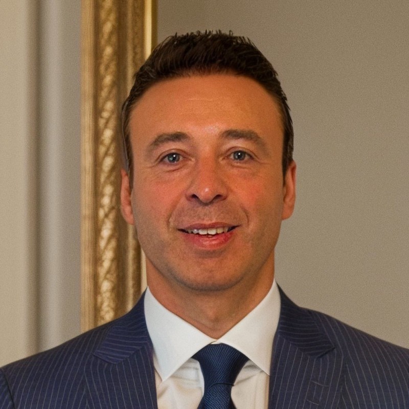 Photo of Fabio; A professional headshot of a middle-aged man with short, styled dark brown hair, blue eyes, and a light complexion. He is smiling warmly, showing his teeth, and is dressed in a formal blue pinstripe suit jacket, a white collared shirt, and a solid dark blue tie. The background is a soft, muted grey with a blurred vertical gold-colored frame visible on the left side.