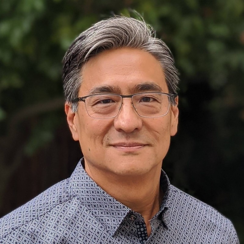 Photo of Danny Yu event speaker; A close-up portrait of an asian man with short, dark brown hair and light eyes, wearing thin-rimmed oval glasses and a dark shirt, set against a softly blurred indoor background.