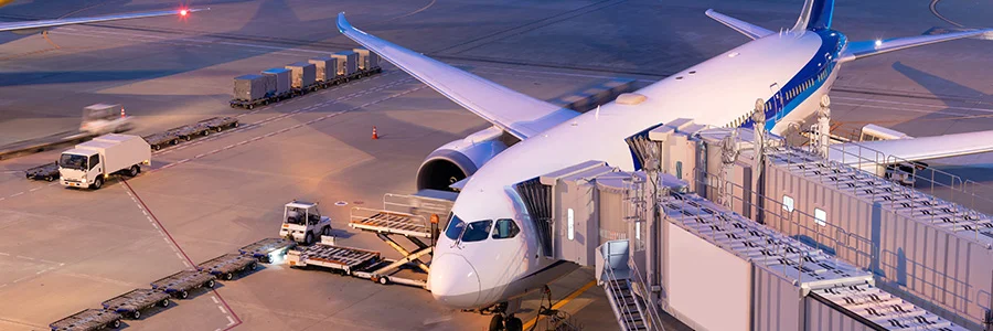 A commercial airplace at an airport gate during ground operations, with baggage carts, service vehicles, and a jet bridge connected