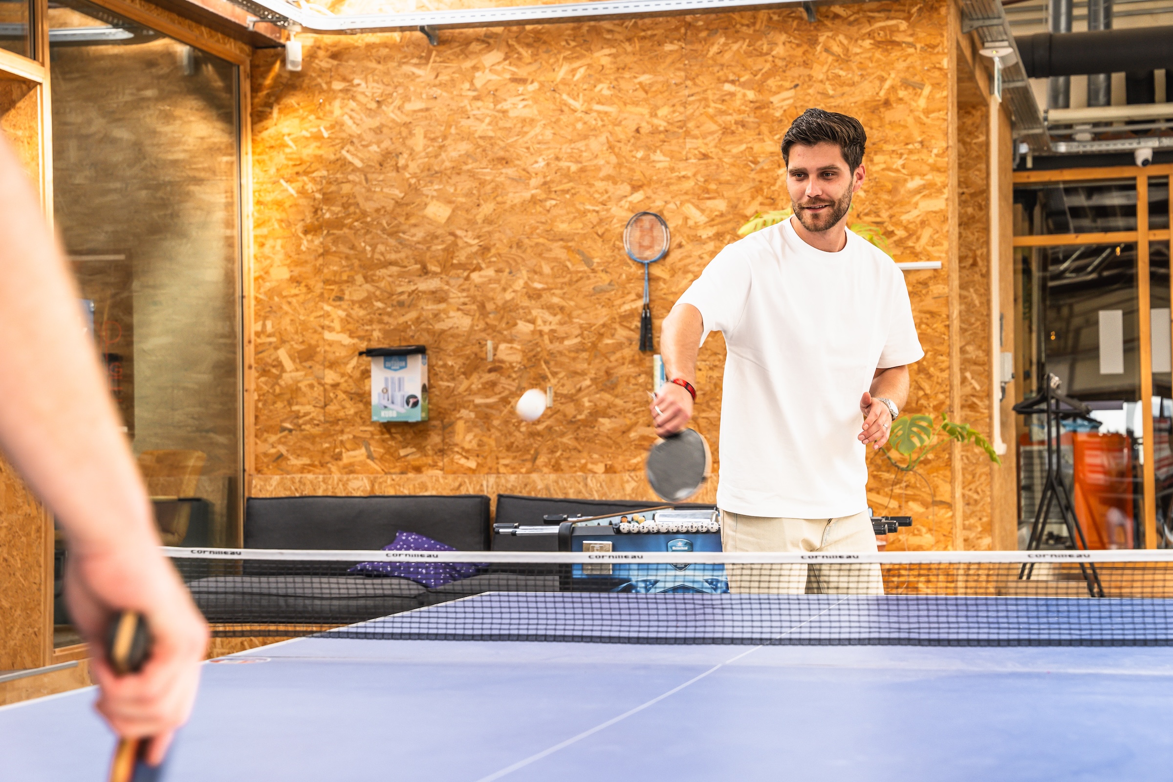 Man in white shirt playing pingpong in a modern indoor recreational room with wood-paneled walls.