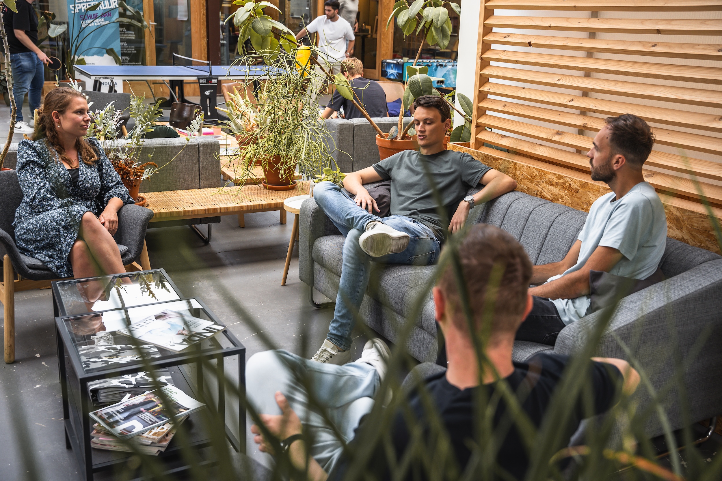 Vier mensen zitten ontspannen in een moderne lounge met planten, een glazen koffietafel en een tafeltennistafel op de achtergrond.