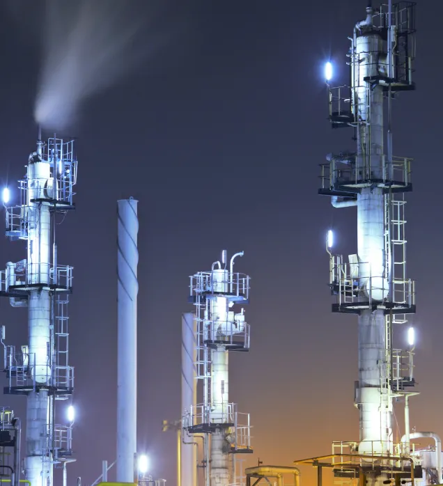 Industrial gas processing plant with illuminated tall towers and smoke venting against a night sky.