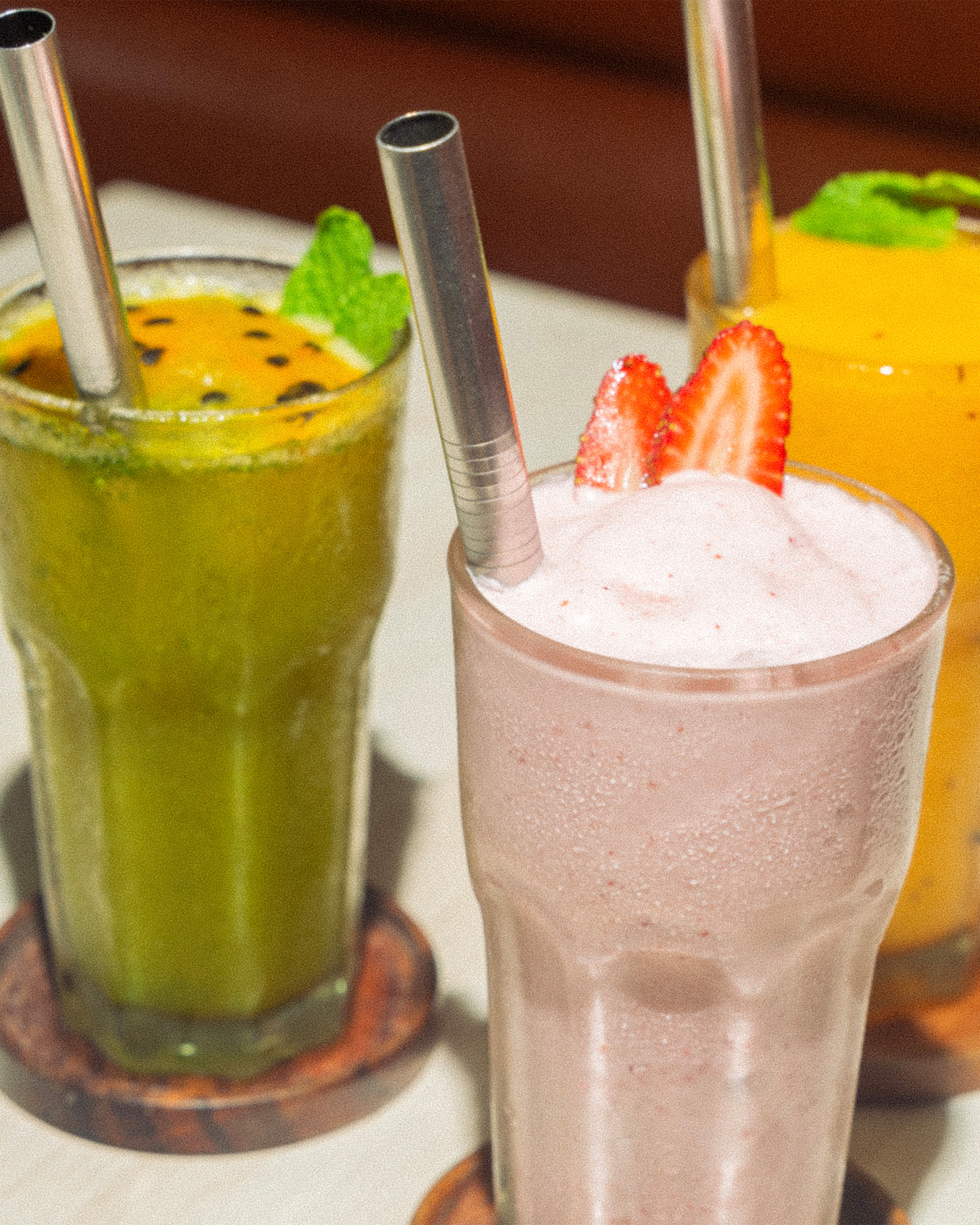 Trois smoothies colorés dans des verres avec paille en acier, garnis de fraises et de feuilles de menthe.