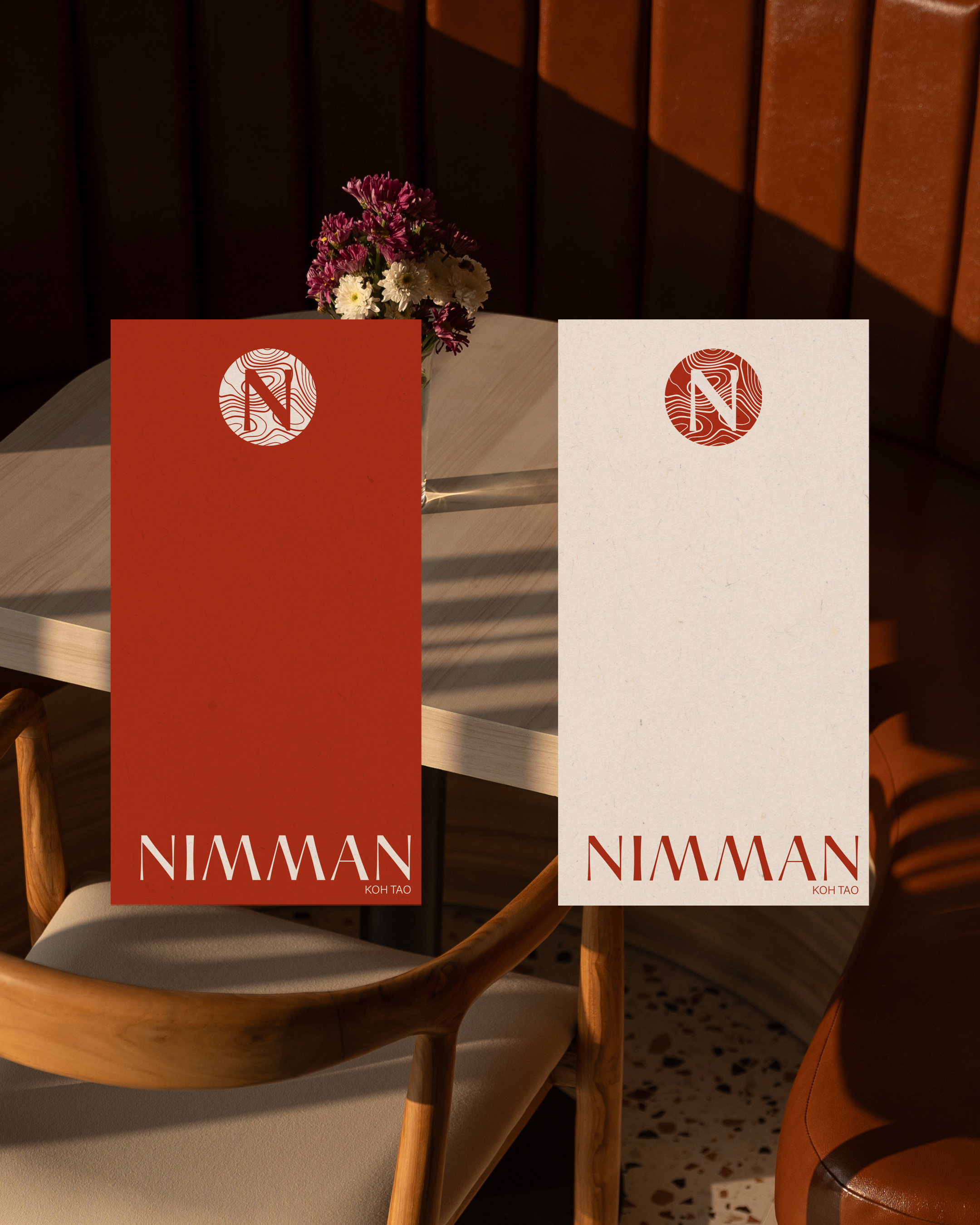 Deux menus élégants posés sur une table en bois dans un restaurant, un rouge foncé et un beige, avec le logo circulaire stylisé et le texte "NIMMAN KOH TAO".