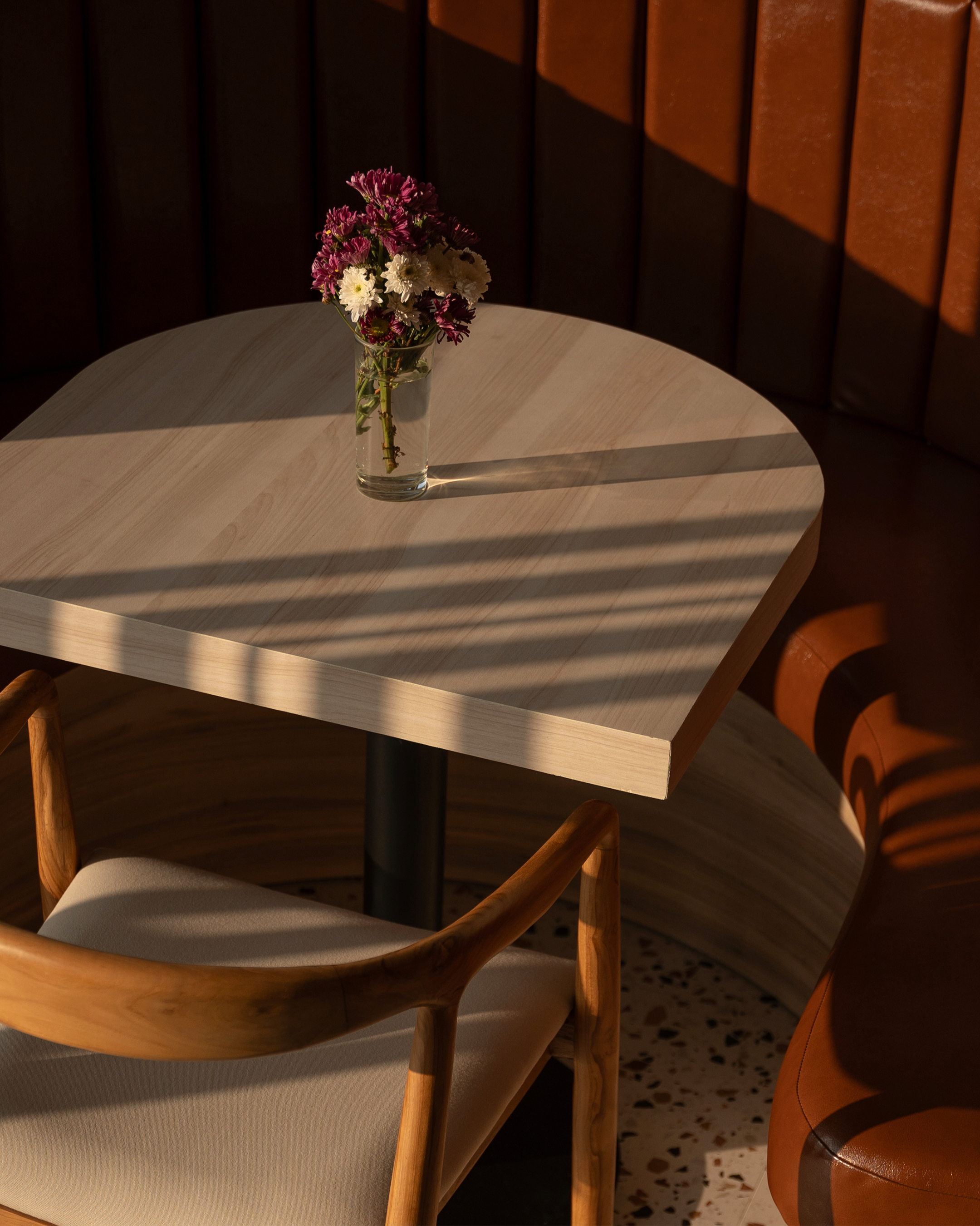 Petite table en bois avec un vase de fleurs mauves et blanches, entourée d'une chaise en bois clair et banquette en cuir brun, éclairée par la lumière du soleil.