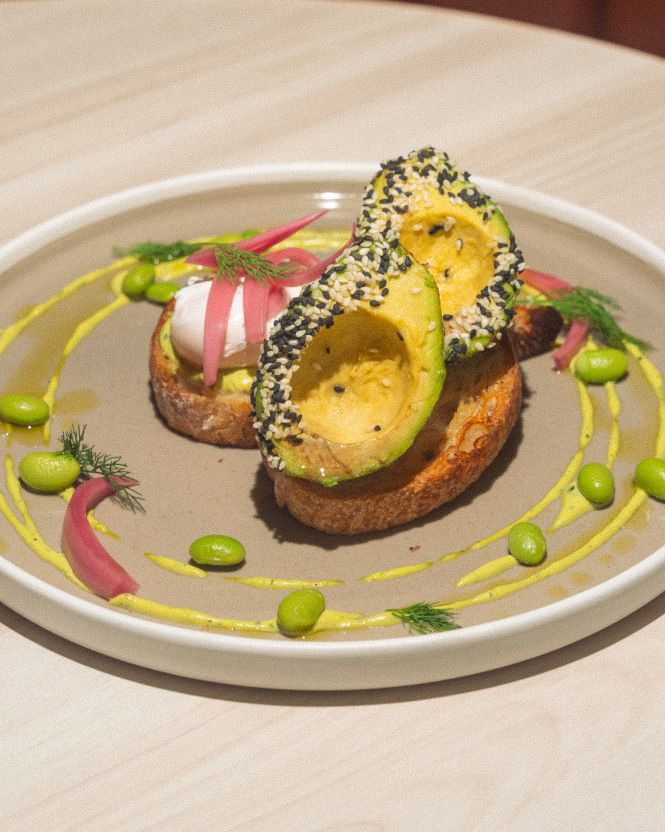 Deux tranches de pain grillé garnies d'avocat aux graines de sésame, œuf poché et oignon rouge, décorées de sauce verte, edamames et herbes fraîches sur une assiette grise.