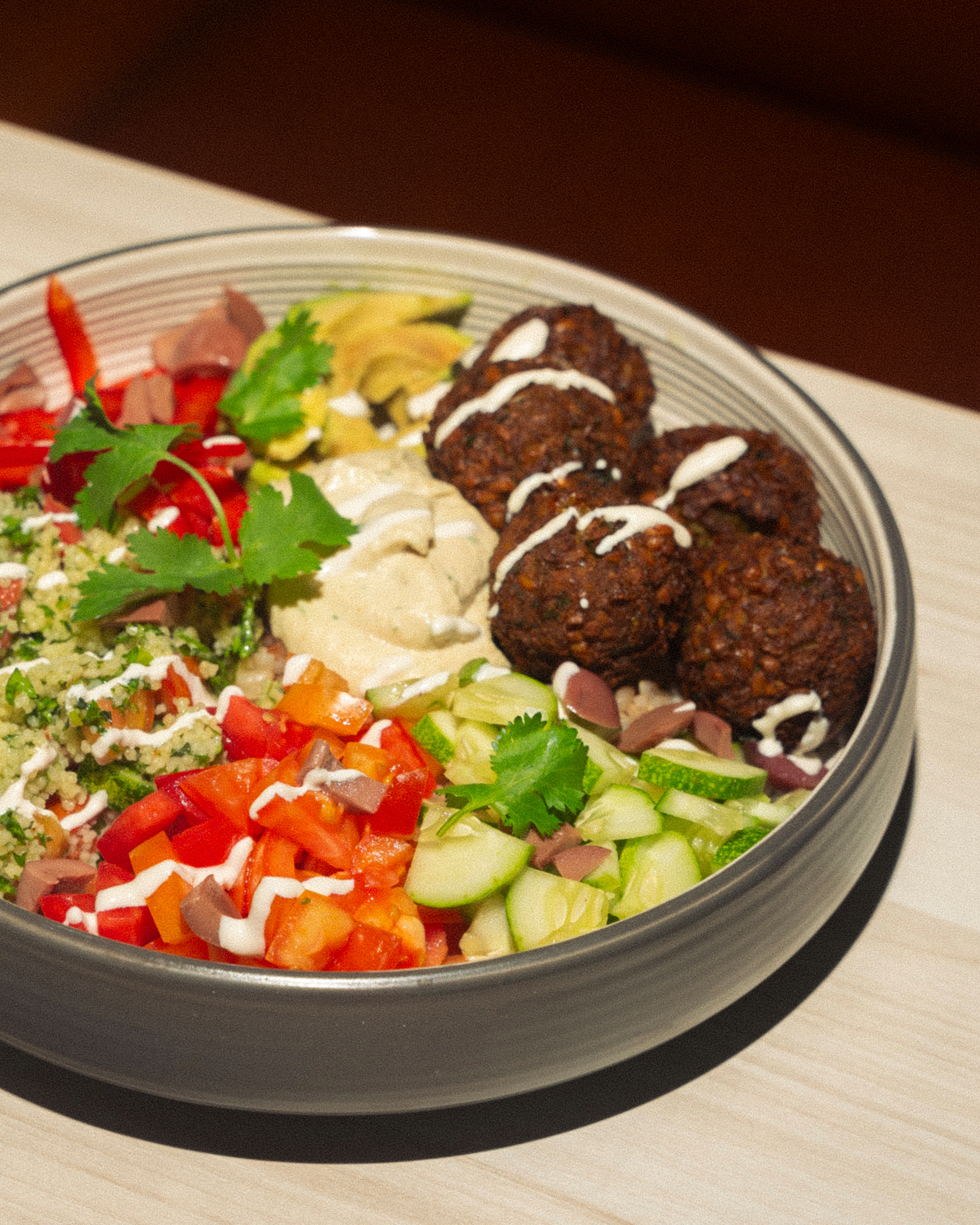 Bol de falafels avec houmous, concombres, tomates, poivrons rouges, olives, persil, et sauce blanche, posé sur une table en bois.