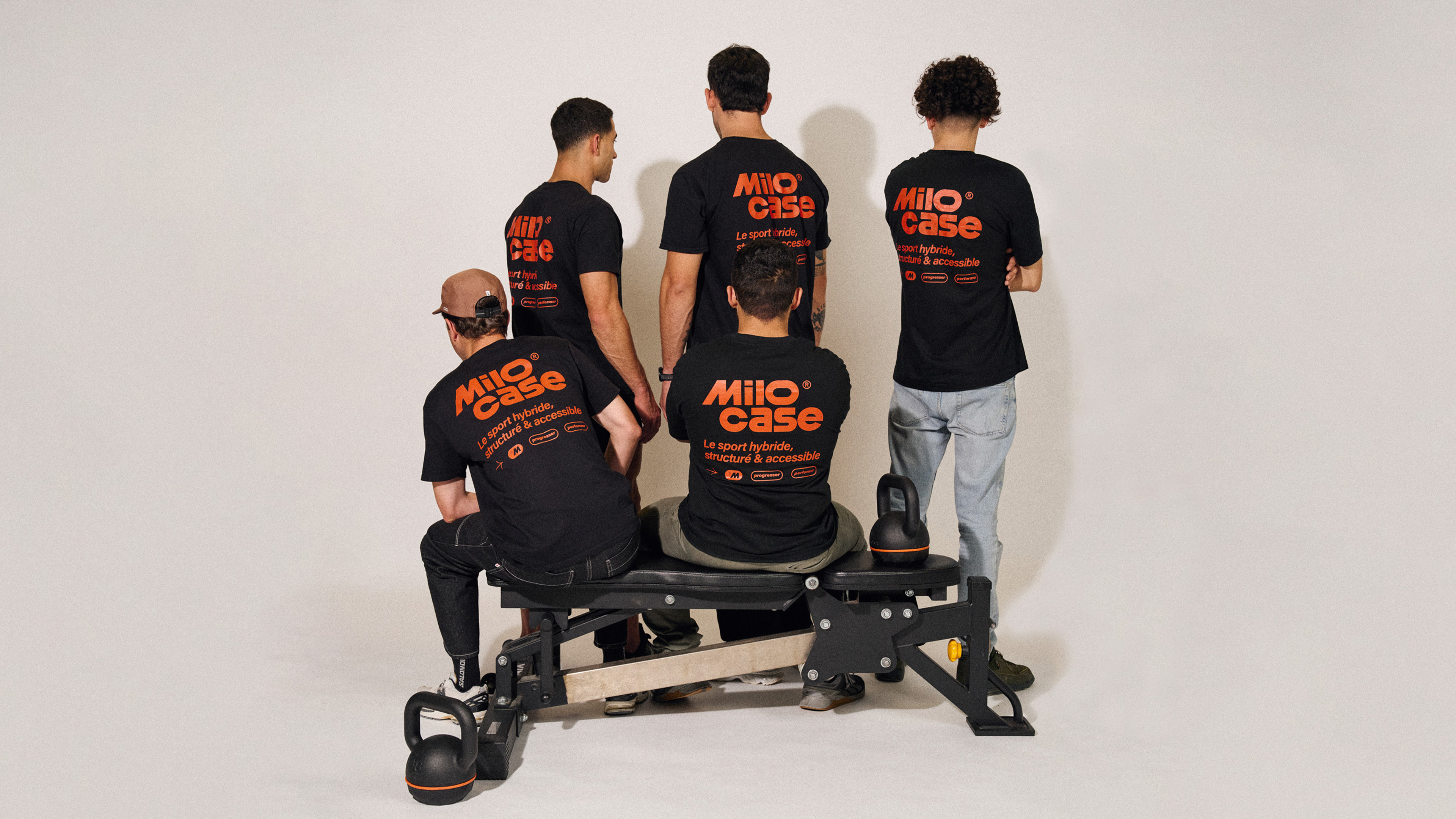 Groupe de cinq hommes vus de dos portant des t-shirts noirs avec le texte orange « Milo case Le sport hybride, structuré & accessible », autour d’un banc de musculation avec deux kettlebells au sol.