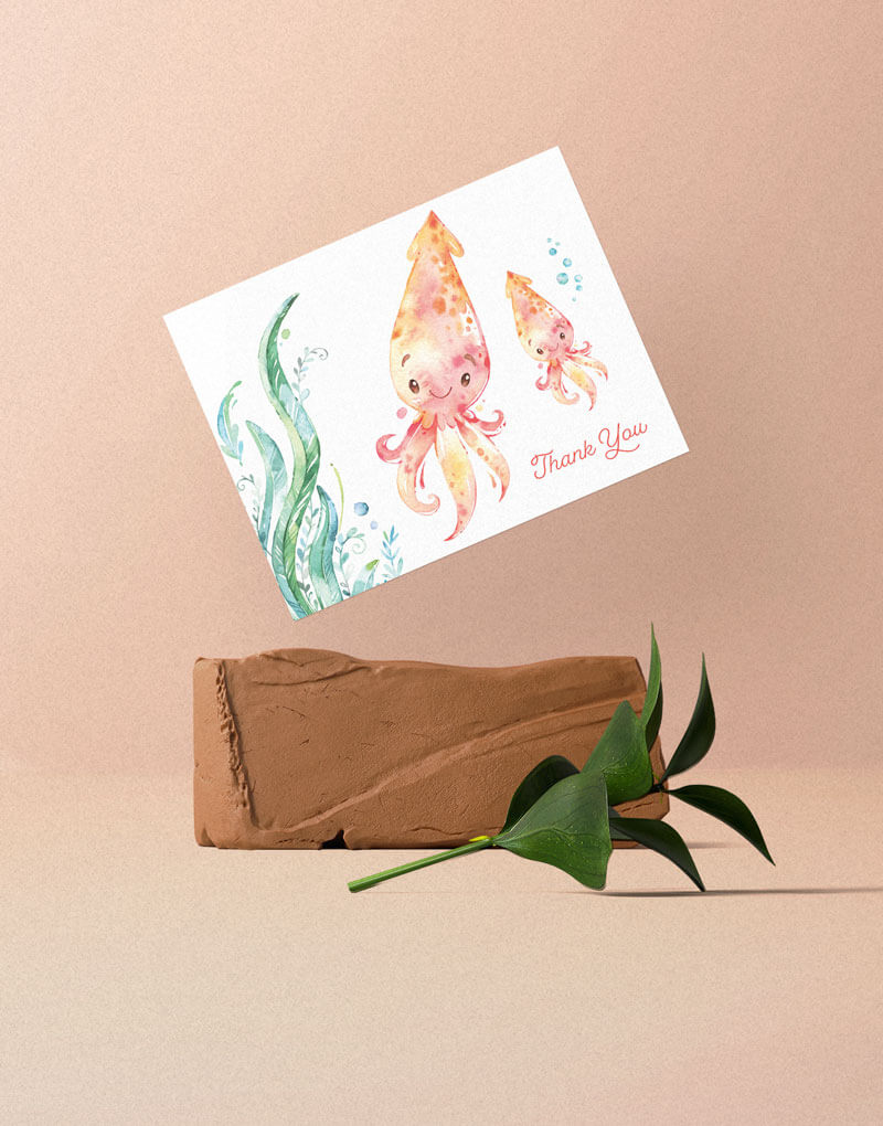 A greeting card with mother and baby squid illustration. Placed with the angle on top of an orange rock with a green branch in front of it.